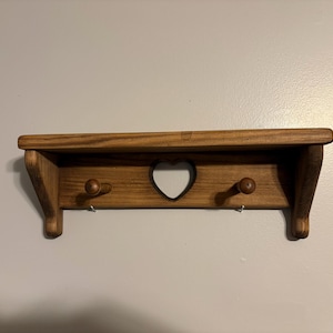 May include: Wooden wall shelf with a heart-shaped cutout and two wooden pegs. The shelf is stained a warm brown color and has a small shelf underneath. The shelf is designed for hanging items and adds a rustic touch to any room.