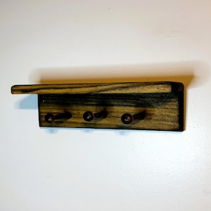 May include: A wooden wall-mounted shelf with three pegs for hanging items. The shelf has a dark, stained finish with visible wood grain. The pegs are round and protrude from the bottom of the shelf. The shelf has a small, overhanging top.