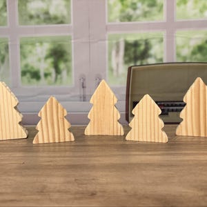 May include: Five wooden Christmas tree ornaments in a row. The trees are light-colored, with a natural wood grain. The background includes a window and a vintage radio.