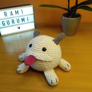 Op de afbeelding: Een handgemaakt wit gehaakt wezen met grijze oren, zwarte ogen en een roze tong. Het pluche speelgoed ligt op een houten oppervlak. Een lichtbak met de tekst "RAMI GURUMI" staat op de achtergrond.