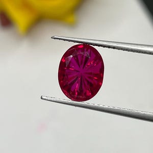Puede incluir: Una gema de rubí de corte ovalado con un tono rojo intenso y una superficie facetada, sostenida firmemente por unas pinzas de metal plateado. La piedra preciosa muestra un patrón radial distintivo de reflejos de luz en su superficie.