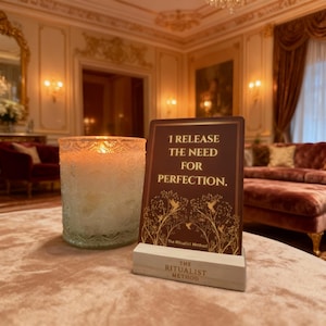 Puede incluir: Una vela encendida en un tarro de cristal decorativo y una tarjeta con el texto "I RELEASE THE NEED FOR PERFECTION" sobre un soporte de madera. La tarjeta tiene un fondo marrón con detalles dorados.