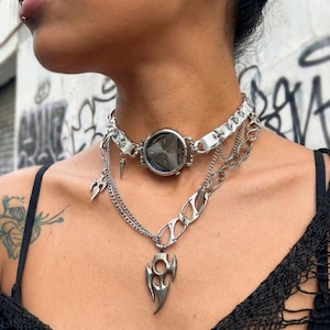 May include: A silver-toned choker necklace with a large, round, dark-colored center piece. The necklace features multiple chains and decorative pendants, including a spike and a stylized flame design. The jewelry is worn around the neck.
