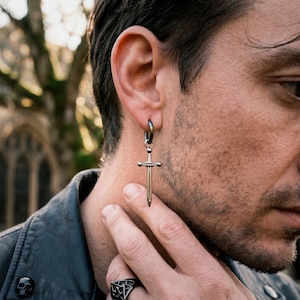 May include: A silver sword pendant earring suspended from a black hoop earring. The earring is worn by a person in a black leather jacket. A black ring with a geometric design is visible on the person's finger.