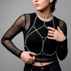 May include: Silver-colored body chain featuring a choker-style necklace connected to a series of chains and rings that create a chest harness design. The jewelry is worn over a black long-sleeved top.