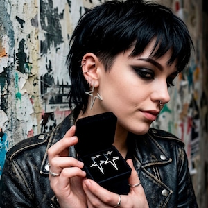 May include: A pair of silver star-shaped earrings, one worn and one in a black jewelry box. The earrings are a minimalist design, with a five-pointed star outline. The person is wearing a black leather jacket.