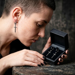 May include: Black sword-shaped earrings with hoop closures. One earring is worn on the ear, and a matching pair is displayed in a black jewelry box. The earrings have a gothic or edgy aesthetic.