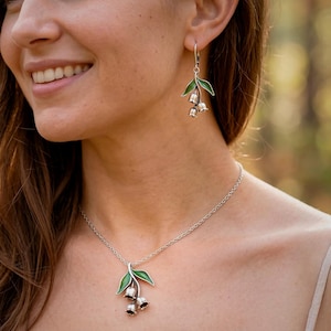 May include: Silver necklace and earrings set. The pendant and earrings feature a green leaf design with three bell-shaped flowers. The jewelry is worn by a person. The background is blurred, suggesting an outdoor setting.