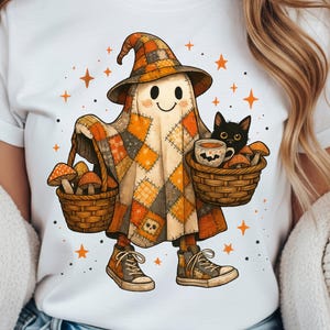 Pode incluir: Camiseta branca com um personagem de desenho animado fantasma em uma roupa de retalhos e chapéu de bruxa, carregando cestas de cogumelos e um gato preto. O fantasma está sorrindo, com estrelas laranja espalhadas pelo design. O fantasma está usando tênis marrons e laranja.