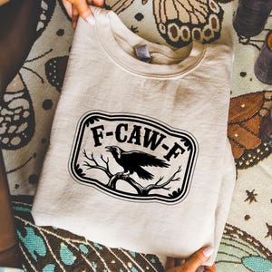 May include: A beige sweatshirt with a black and white graphic. The graphic features the text "F-CAW-F" in a black, decorative frame, with a black raven perched on a branch. The sweatshirt is folded.