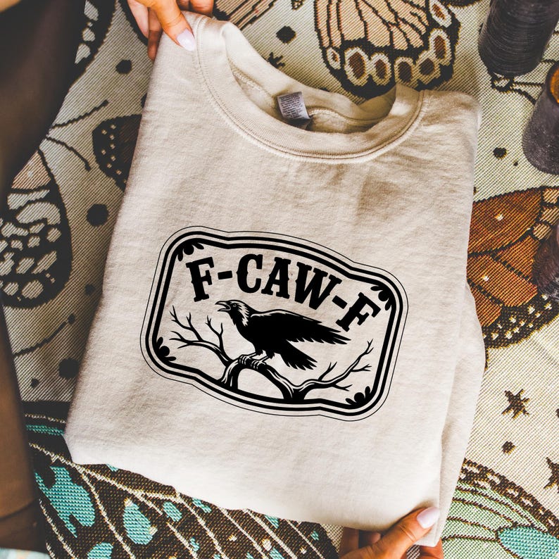 May include: A beige sweatshirt with a black and white graphic. The graphic features the text "F-CAW-F" in a black, decorative frame, with a black raven perched on a branch. The sweatshirt is folded.