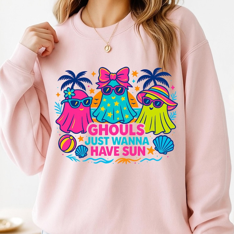 May include: Pink sweatshirt featuring a graphic of three cartoon ghosts wearing sunglasses and beach attire. The text "GHOULS JUST WANNA HAVE SUN" is printed below the ghosts. The design includes palm trees, stars, and a beach ball.