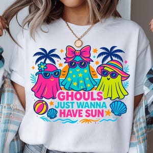 May include: White t-shirt featuring a colorful graphic of three cartoon ghosts wearing sunglasses and beach attire. The text "GHOULS JUST WANNA HAVE SUN" is printed below the ghosts, with beach-themed illustrations.
