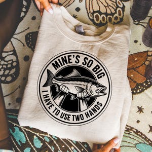 May include: A beige sweatshirt with a black and white graphic of a fish held by two hands, encircled by the text "MINE'S SO BIG I HAVE TO USE TWO HANDS". The sweatshirt is folded.