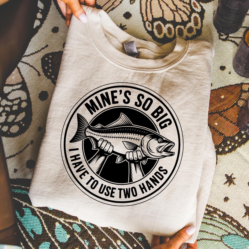 May include: A beige sweatshirt with a black and white graphic of a fish held by two hands, encircled by the text "MINE'S SO BIG I HAVE TO USE TWO HANDS". The sweatshirt is folded.