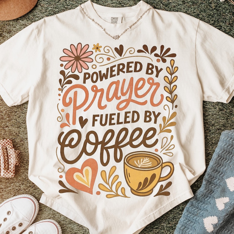 May include: Cream-colored t-shirt with the text "POWERED BY Prayer FUELED BY Coffee" in a decorative design. The design includes floral and coffee-related illustrations in shades of brown, orange, and yellow. A casual, comfortable tee.