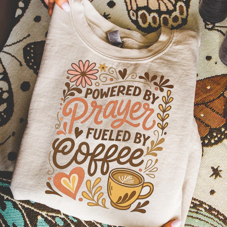 May include: A beige sweatshirt with the text "POWERED BY Prayer FUELED BY Coffee" in a decorative font. The design includes floral and coffee-themed illustrations in shades of brown, peach, and gold. The sweatshirt is folded.