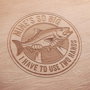 May include: A circular wooden carving with the text "MINE'S SO BIG I HAVE TO USE TWO HANDS" surrounding a fish held by two hands. The fish has an open mouth and detailed scales. The background is a light brown wood grain.