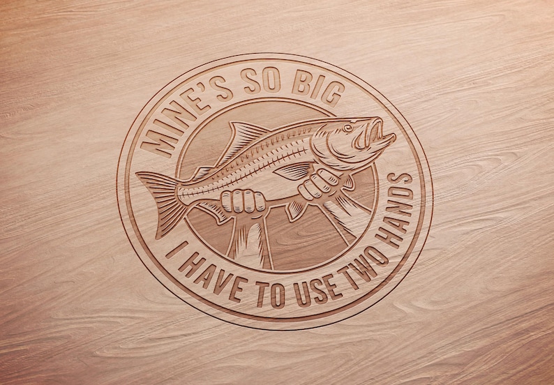 May include: A circular wooden carving with the text "MINE'S SO BIG I HAVE TO USE TWO HANDS" surrounding a fish held by two hands. The fish has an open mouth and detailed scales. The background is a light brown wood grain.