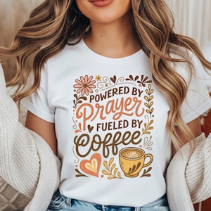 May include: White t-shirt with the text "Powered by Prayer Fueled by Coffee." The design includes floral and heart accents, a coffee cup, and the words in brown and orange colors.