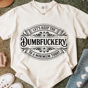 Puede incluir: Camiseta de color crema con un gr&aacute;fico negro que dice "LET'S KEEP THE DUMBFUCKERTY TO A MINIMUM TODAY." La camiseta tiene cuello redondo y mangas cortas. Se ve un collar plateado en el escote. La camiseta est&aacute; doblada sobre una superficie texturizada.