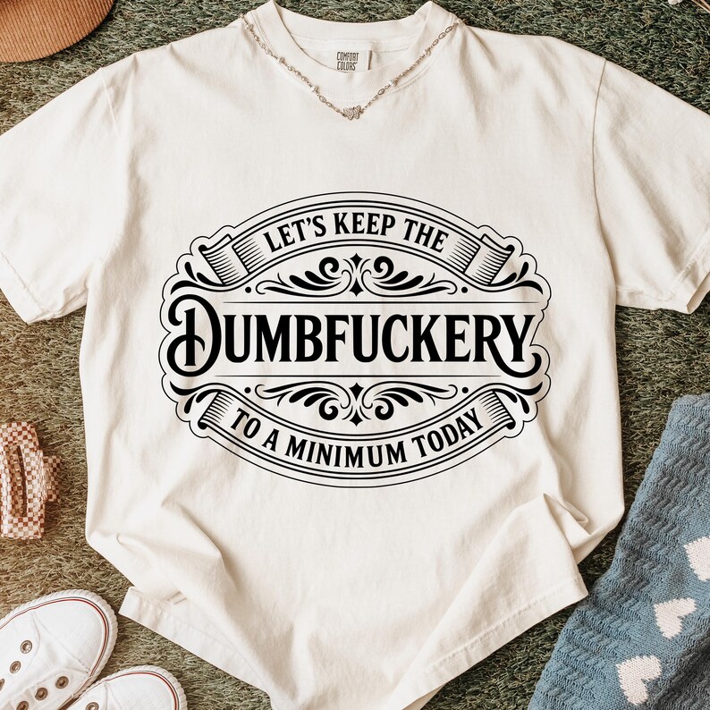 Puede incluir: Camiseta de color crema con un gr&aacute;fico negro que dice "LET'S KEEP THE DUMBFUCKERTY TO A MINIMUM TODAY." La camiseta tiene cuello redondo y mangas cortas. Se ve un collar plateado en el escote. La camiseta est&aacute; doblada sobre una superficie texturizada.