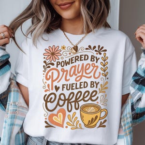 May include: White t-shirt with the text "POWERED BY Prayer FUELED BY Coffee" in brown and orange. The design includes floral and coffee cup illustrations. The shirt is a casual wear item.