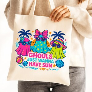May include: A cream-colored tote bag featuring a colorful graphic of three ghost characters wearing sunglasses and beach attire. The text "Ghouls Just Wanna Have Sun" is printed below the ghosts, with palm trees and beach elements.
