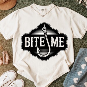 May include: Cream-colored t-shirt with a black and white graphic of a fishing hook and the words "BITE ME". The shirt has a V-neck and short sleeves. A necklace is visible at the neckline.