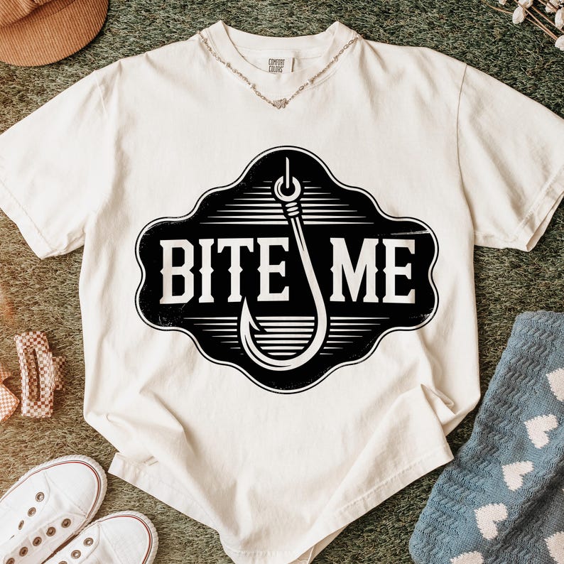 May include: Cream-colored t-shirt with a black and white graphic of a fishing hook and the words "BITE ME". The shirt has a V-neck and short sleeves. A necklace is visible at the neckline.