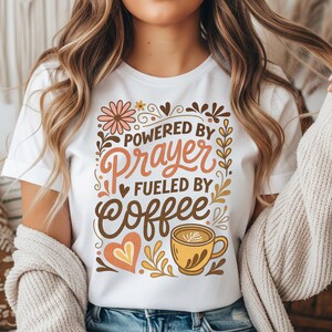 May include: White t-shirt with the text "Powered by Prayer Fueled by Coffee" in a decorative design. The design includes a coffee cup, flowers, and hearts in shades of brown, orange, and pink.