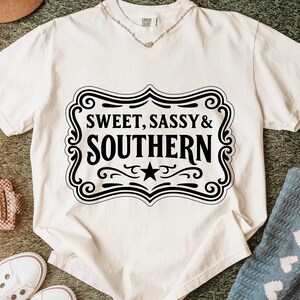 May include: Off-white t-shirt with the text "SWEET, SASSY & SOUTHERN" in a black decorative frame. The shirt has short sleeves and a crew neck. A silver necklace is visible at the neckline. The shirt is on a green surface.
