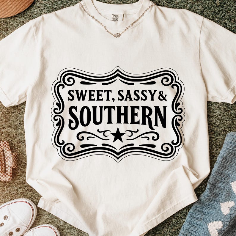 May include: Off-white t-shirt with the text "SWEET, SASSY & SOUTHERN" in a black decorative frame. The shirt has short sleeves and a crew neck. A silver necklace is visible at the neckline. The shirt is on a green surface.