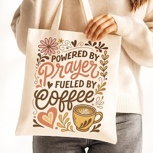 May include: Cream-colored tote bag with the text "Powered by Prayer Fueled by Coffee." The design includes floral and heart motifs, and a coffee cup illustration. The bag has cream-colored handles.