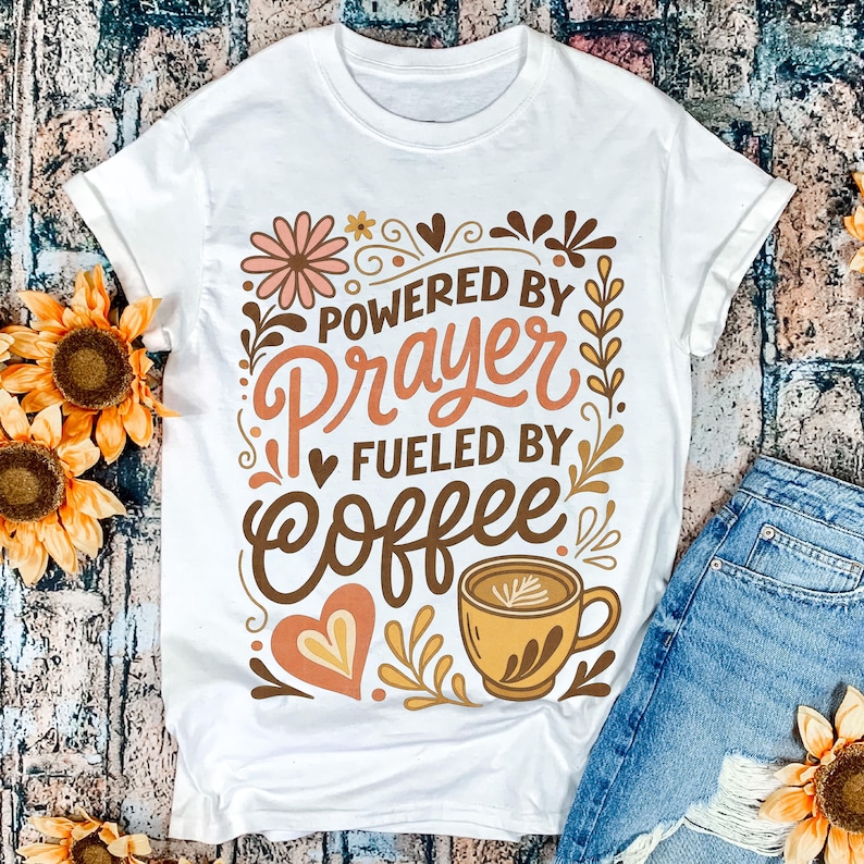 May include: White t-shirt with the text "POWERED BY PRAYER FUELED BY COFFEE" in brown, orange, and peach colors. The design includes a coffee cup, hearts, and floral elements. The shirt is paired with blue denim shorts.