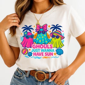 May include: White t-shirt featuring a colorful graphic of three cartoon ghosts wearing sunglasses and beach attire. The text "GHOULS JUST WANNA HAVE SUN" is below the ghosts. The design includes palm trees, stars, a beach ball, and a seashell.