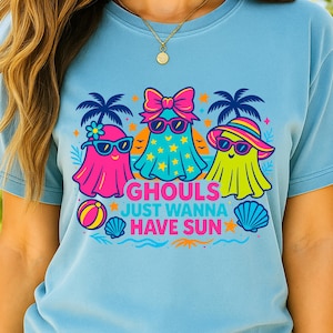 May include: Light blue t-shirt featuring a graphic of three cartoon ghosts wearing sunglasses and beach attire. The text "GHOULS JUST WANNA HAVE SUN" is printed below the ghosts, along with beach-themed illustrations.