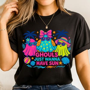 May include: Black t-shirt featuring a colorful graphic of three cartoon ghosts wearing sunglasses and beach attire. The text "GHOULS JUST WANNA HAVE SUN" is displayed below the ghosts, with stars and beach-themed elements.