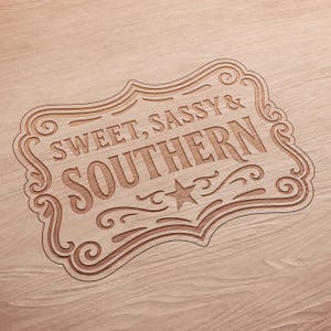 May include: A wooden sign with the words "SWEET, SASSY & SOUTHERN" carved into it. The sign has a decorative border with swirls and a star. The sign is light brown and sits on a light brown wooden surface.