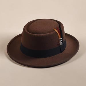 May include: A brown felt pork pie hat featuring a flat crown and a narrow, upturned brim. The hat is accented with a wide black grosgrain ribbon band and a small decorative cluster of orange, brown, and black spotted feathers tucked into the side.