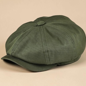 May include: An olive green newsboy cap featuring an eight-panel construction with a central fabric-covered button on the crown and a short, curved brim. The hat is crafted from a solid-colored, textured fabric with visible stitching along the seams.