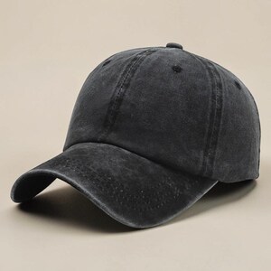 May include: A charcoal black baseball cap with a washed, distressed finish. The cap features a six-panel construction with a curved brim, a button at the top centre, and stitched eyelets for ventilation, presenting a worn-in aesthetic.
