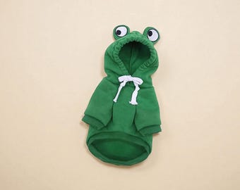 May include: A green pet hoodie with a frog design. The hoodie has a hood with cartoon eyes and a white drawstring. The garment is designed to fit a small dog or cat.