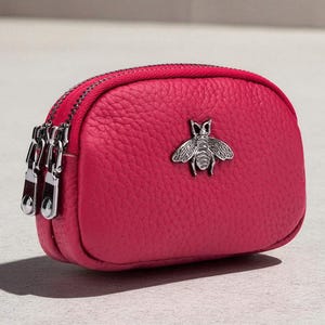 Puede incluir: Un pequeño monedero de cuero color frambuesa con un emblema de abeja plateada. El bolso tiene una superficie texturizada, una cremallera doble y una forma redondeada. Los herrajes son plateados.