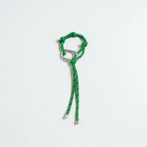 Peut inclure: Un porte-clés en corde verte et noire avec un mousqueton argenté. Le porte-clés est noué en forme circulaire avec deux brins pendants, chacun terminé par une extrémité métallique argentée.