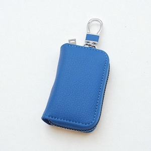 Puede incluir: Un estuche para llaves de cuero azul con un cierre y cremallera de metal plateado. El estuche para llaves es rectangular con bordes redondeados y una superficie texturizada. La cremallera es negra.