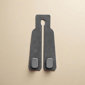 May include: Two gray leather car seat belt covers with a metal accent. The covers have a stitched border and a slot at the top for hanging. The metal accents are rectangular and placed near the bottom.