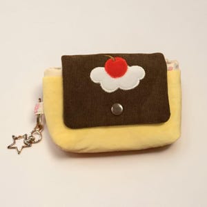 May include: A small yellow pouch with a brown corduroy flap featuring an embroidered white cloud and a red cherry. The pouch includes a silver metal snap closure and a silver star-shaped charm attached to a side loop with a small metal ring.
