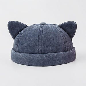 May include: A dark blue denim docker cap featuring two prominent, upright cat-like ears attached to the crown. The hat has a rounded, six-panel construction with a thick, folded brim and a small fabric button at the very top center.