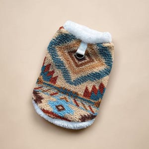 Pode incluir: Um suéter para cães com um padrão geométrico em tons de castanho, azul e vermelho. O suéter tem uma gola branca e fofa e um pequeno laço com um anel de metal. O design geral é reminiscente do estilo do sudoeste.